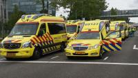 Meerdere gele ambulances met rode en blauwe strepen staan op een rij geparkeerd op straat. Sommige ambulancedeuren staan open. De achtergrond bestaat uit bomen, gebouwen en straatnaamborden.