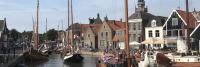 Een kanaal vol met traditionele Nederlandse zeilboten en historische gebouwen in een klein stadje. Mensen lopen en zitten aan het water. De scène omvat een bewolkte lucht, een klokkentoren en zitplaatsen buiten in een café.