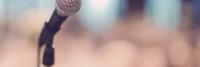 Close-up van een microfoon tegen een zacht onscherpe achtergrond, mogelijk op een podium, waarbij de metaalachtige textuur en de bereidheid voor een spreker of artiest worden benadrukt.