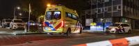 Een gele ambulance staat 's nachts op straat geparkeerd, met zwaailichten. In de omgeving zijn politieauto's aanwezig en een deel van het gebied is afgezet met een oranje en wit afzetlint. De achtergrond bestaat uit een gebouw met meerdere verdiepingen en