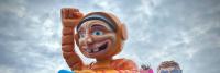 Een grote, kleurrijke praalwagen met een cartoonachtige figuur in een oranje gestreept pak, opgeheven arm en grote ogen aan de voorkant. Een andere kleinere praalwagen met een personage dat een stervormige bril draagt, is gedeeltelijk zichtbaar erachter o