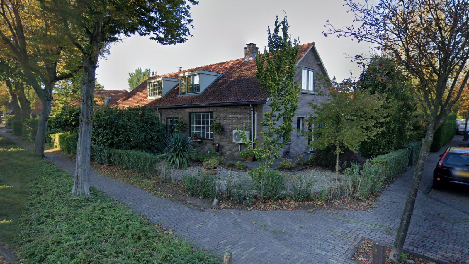 Traditioneel bakstenen huis met puntdak, omgeven door groen en een geplaveide straat in een woonwijk.