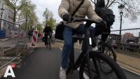 Een fietser rijdt op een fietspad langs een kanaal, met andere fietsers en voetgangers in de buurt. Er zijn wegwerkzaamhedenborden zichtbaar die mogelijke omwegen of wegwerkzaamheden aangeven.