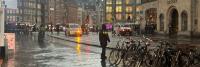 Doorweekte straat in een stad met reflecties van voertuiglichten op de natte stoep, fietsen geparkeerd aan de zijkant en voetgangers met paraplu's.