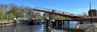 Een moderne voetgangersbrug met een opvallend hoekig ontwerp steekt op een zonnige dag een kalm water over, met helderblauwe luchten en weelderig groen op de achtergrond.