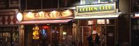 Nachtelijk zicht op een stedelijke straat met verlichte borden van 'café cuba' en 'cotton club' boven etablissementen, met voorbijlopende mensen en geparkeerde fietsen.