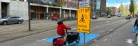 Een persoon in een rood jasje rijdt op een bakfiets op een aangewezen fietspad dat blauw is gemarkeerd. Er staat een geel bord met een waarschuwingsbericht in het Nederlands dat zich vertaalt naar 'Let op: fietsers op de rijbaan'. Links staat een grijze a