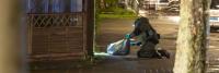 Een politieagent onderzoekt 's nachts buiten een plaats delict, gehurkt met een zaklamp naast een voorwerp op de grond, met geparkeerde auto's en woongebouwen op de achtergrond.