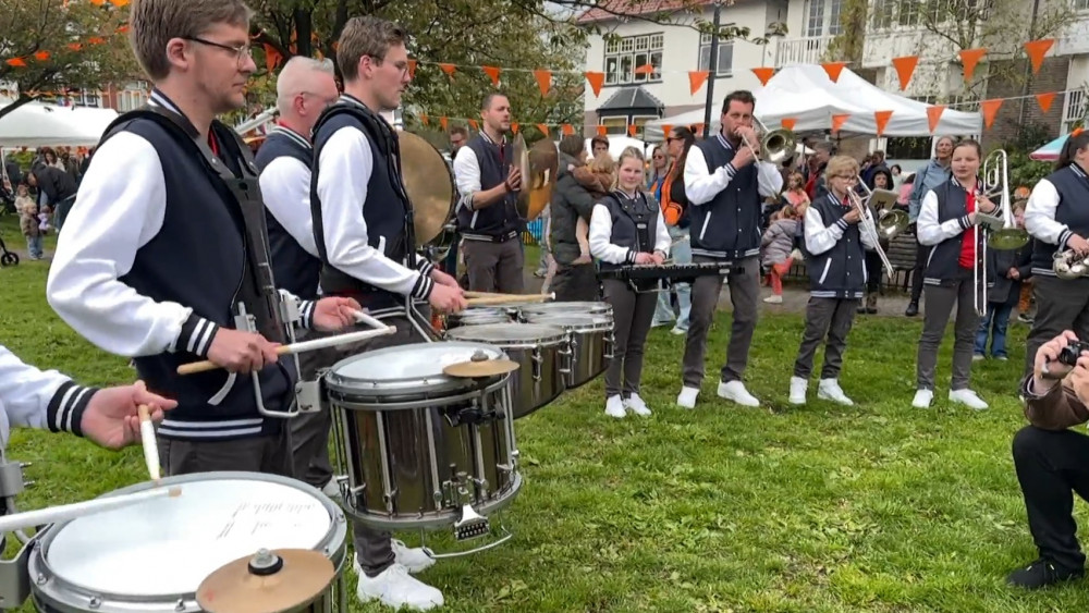 Een groep muzikanten, gekleed in traditionele vesten en witte shirts, speelt drums en een fluit op een openluchtfestival versierd met oranje en witte vlaggetjes.