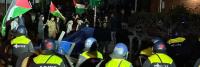Een nachtelijke scène van een protest waarbij demonstranten Palestijnse vlaggen vasthouden en tegenover een rij politieagenten in oproeruitrusting staan. de setting is stedelijk, verlicht door kunstlicht.