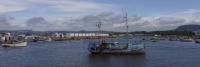 Panoramisch zicht op een haven met diverse boten, waaronder een prominent versierde boot met kleurrijke vlaggen en sculpturen, tegen een achtergrond van lage heuvels en een bewolkte lucht.