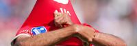 Een voetballer die een rode trui draagt, trekt het shirt over zijn hoofd, waardoor zijn gezicht gedeeltelijk wordt verborgen. Zijn rechterarm is gebogen terwijl hij het shirt optilt, en op de mouw is een blauw-witte sponsorpatch zichtbaar.