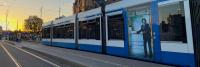 Een blauw-witte tram wordt tijdens zonsondergang gestopt bij een station. Op de tram staat een advertentie met een vrouw in formele kledij die naast een receptie staat. Op het perron zijn mensen zichtbaar en op de achtergrond zijn gebouwen te zien.