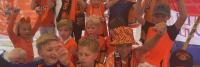 Een groep kinderen, gekleed in oranje kleding en accessoires, juicht enthousiast en steekt hun hand op. Sommigen dragen hoeden en er is een vlag op de achtergrond. De setting lijkt zich onder een tent te bevinden.
