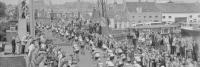 Zwart-witfoto van een druk straatbeeld met een grote groep wielrenners in een wedstrijd, mogelijk een beroepsevenement. Toeschouwers staan langs beide zijden van de smalle straat en sommigen staan op een brug en nabijgelegen daken. Gebouwen en een boot op