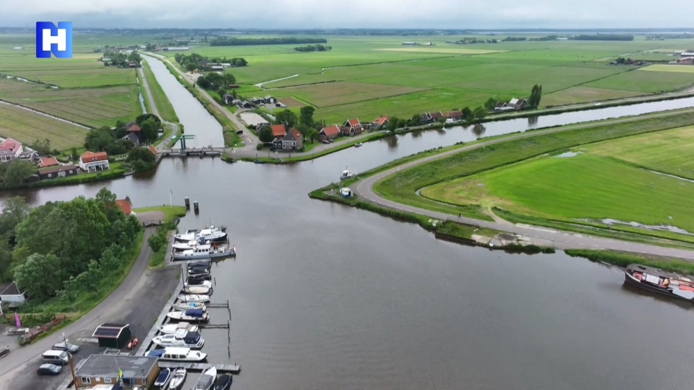 Luchtfoto van een landelijke jachthaven met verschillende boten aangemeerd, omgeven door weelderige groene velden en elkaar kruisende grachten. Kleine huizen en gebouwen liggen verspreid langs de waterkant en in de verte is een kleine brug zichtbaar. Bewo