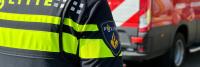 Een close-up van de achterkant van een politieagent die een uniform draagt met de tekst 'Politie' erop en die naast een rood hulpverleningsvoertuig staat. Op de schouderpatch van de officier staat een fakkelsymbool.