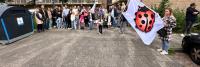 Een groep mensen, inclusief volwassenen en kinderen, verzamelt zich op een verhard terrein nabij een appartementengebouw. Rechts staat een vrouw met een grote witte vlag met daarop een rood en zwart lieveheersbeestje. Links staat een grote zwart-blauwe ba