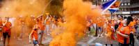 Buiten verzamelt zich een groep mensen, gekleed in oranje kleding en zwaaiend met oranje rookfakkels. Sommigen houden vlaggen en spandoeken vast. De scène is feestelijk en energiek, met gebouwen en bomen zichtbaar op de achtergrond.