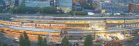 Een luchtfoto van een druk stedelijk treinstation. Er zijn meerdere treinperrons zichtbaar met op sommige sporen treinen. Grenzend aan de perrons ligt een rijbaan met geparkeerde bouwvoertuigen. Rondom het station staan talloze moderne hoogbouwgebouwen.