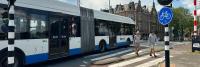 Een wit-blauwe gelede bus rijdt in een stedelijk gebied en passeert een zebrapad. Er lopen twee mensen over het zebrapad. Fietspaden en een fietsbord zijn zichtbaar, samen met historische gebouwen op de achtergrond.