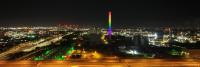 Een panoramisch uitzicht op een industriegebied 's nachts met gebouwen en constructies verlicht door verschillende lichten. Centraal in de afbeelding staat een hoge schoorsteen, met een verloop van regenboogkleuren. De achtergrond toont een stadsgezicht m