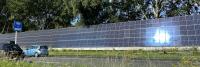 Een lange reeks zonnepanelen ligt langs de kant van een weg op een zonnige dag. Er staan drie auto's vlakbij geparkeerd en aan de linkerkant is een blauw bord met gele letters zichtbaar. Op de achtergrond wordt het gebied begrensd door bomen.