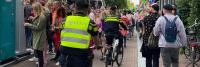 Een groep mensen, sommigen gekleed in kleurrijke kleding, loopt door een drukke straat naast geparkeerde fietsen. Er worden twee politieagenten in gele en zwarte uniformen gezien op de fiets, waarbij ze zichtbaar aanwezig blijven. Daarnaast zijn aan de li