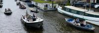 Een kanaal met verschillende kleine boten gevuld met mensen die op hun gemak over het water varen. Aan de rechterzijde ligt een woonboot met groendak. Gebouwen, auto's en groen langs de randen van het kanaal. Mensen zitten op de boten en genieten van het 