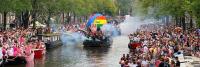 Een levendige menigte staat langs de gracht in een stad voor een parade. Door het water bewegen boten gevuld met mensen, waaronder één met een grote regenboogbol en een opgeheven handfiguur. Rookeffecten dragen bij aan de feestelijke sfeer terwijl bomen a