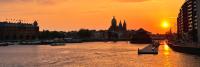 De afbeelding toont een stadsgezicht aan het water bij zonsondergang. De lucht is oranje en de zon staat net boven de horizon. Er zijn boten op het water, gebouwen langs de kust en een kathedraal in silhouet met torenspitsen op de achtergrond.