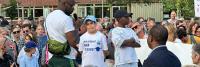 Buiten verzamelt zich een menigte mensen. Een persoon met een microfoon spreekt de groep toe, terwijl twee mensen in witte T-shirts naast elkaar staan. Sommige personen op de achtergrond houden borden vast. Op de achtergrond zijn bomen en gebouwen zichtba