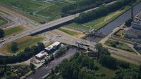 Luchtfoto van een snelwegviaduct dat een kanaal eronder kruist. Rondom het gebied liggen groene velden, gebouwen en bomen. Een kleinere weg loopt parallel aan het kanaal, met meerdere auto&#x27;s zichtbaar op zowel de snelweg als de kleinere weg.
