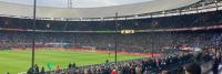 Een vol voetbalstadion met toeschouwers die een wedstrijd bekijken. Het groene veld is in het midden, omringd door tribunes vol fans. Meerdere reclamebanners worden rond het veld getoond en een paar mensen staan vooraan bij het veld.