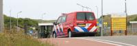 Een rode noodhulpbus met blauwe en witte diagonale strepen staat geparkeerd bij de ingang van een faciliteit. De faciliteit is gemarkeerd met een geel bord met "Welkom" en andere tekst. Het gebied is omgeven door groen en er zijn verschillende straatlanta