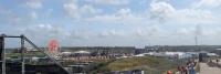 Panoramisch uitzicht op een buitenevenement met verschillende tenten, spandoeken en een reuzenrad op de achtergrond. Mensen lopen langs een pad aan de rechterkant en steigerconstructies zijn zichtbaar aan de linkerkant. De lucht is gedeeltelijk bewolkt.