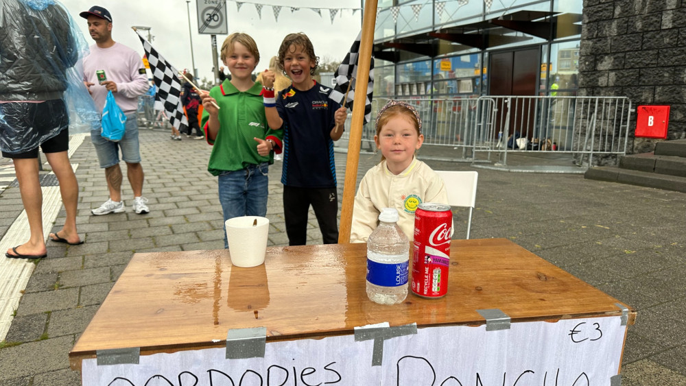 Drie kinderen bij een kleine houten stand met het opschrift “Oordopjes, Poncho, €3” met drankjes erbovenop. Eén kind zit bij de stand en de andere twee houden geblokte vlaggen vast en staan erachter. Ze staan buiten op een nat trottoir bij een gebouw.