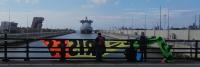 Drie mensen staan bij een metalen reling op een brug en houden een kleurrijke banner voor zich. Beneden is een groot cruiseschip te zien dat aangemeerd ligt in een kanaal, met kranen en industriële structuren zichtbaar aan de horizon. De lucht is gedeelte