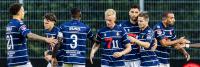 Een groep voetballers in blauw-wit gestreepte shirts loopt over het veld. Sommigen zijn met elkaar in gesprek, terwijl anderen naar voren lopen. Ze bevinden zich in een openluchtstadion met een metalen hek en een rood hart op de achtergrond.