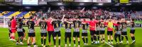 Een voetbalteam in zwart-witte uniformen staat in een rij met hun armen om elkaar heen, tegenover een menigte juichende supporters in een stadion. De stadionstoelen zijn blauw en geel gekleurd en er zijn advertenties op de achtergrond zichtbaar.