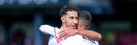 Twee voetballers in witte shirts omhelzen elkaar op het veld. Hun gezichtsuitdrukkingen suggereren een feestelijk moment. De achtergrond is wazig, waardoor de focus op de spelers ligt.