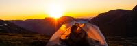 Een persoon zit in een tent bij zonsondergang in een bergachtig gebied. De tent staat op gras en de zon gaat onder achter de bergen. De lucht is levendig met tinten oranje en roze. De persoon in de tent lijkt te ontspannen of te lezen.