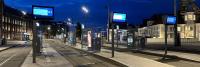 Een modern tramstation onder de avondhemel met blauwe en witte digitale schermen die informatie op elk platform tonen. Verschillende lege platforms en sporen zijn zichtbaar, met gebouwen en straatlantaarns op de achtergrond.