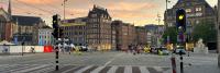 Voetgangersplein bij zonsopgang met verschillende arbeiders in gele hesjes die barrières opzetten. Historische gebouwen en een kerk zijn op de achtergrond te zien. Twee verkeerslichten zijn zichtbaar, één op groen en de andere op rood, samen met een zebra