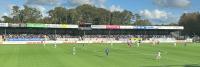 Er is een voetbalwedstrijd gaande in een openluchtstadion. Spelers in blauw-witte uniformen zijn zichtbaar op het veld. Het publiek kijkt toe vanaf de tribune op de achtergrond en reclameborden staan boven op het stadion. Bomen en een bewolkte lucht zijn 