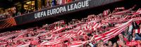 Een grote groep voetbalfans houdt rood-wit gestreepte sjaals van Athletic Club omhoog tijdens een UEFA Europa League-wedstrijd. De menigte zit vol en de fans tonen in koor hun steun.