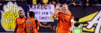 Voetballers in oranje tenues vieren feest op het veld. Een speler steekt zijn hand op. Op de achtergrond is een bord met tekst en diverse logo's zichtbaar. Er zijn toeschouwers aanwezig.