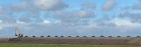 Een gele graafmachine werkt op een aantal heuvels van aarde onder een blauwe lucht met verspreide wolken. De scène speelt zich af in een grasveld, met de graafmachine aan de linkerkant van de afbeelding.