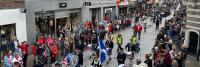 Een parade van een marcherende band trekt door een drukke stadsstraat vol toeschouwers. De bandleden dragen uniformen en hebben vlaggen en instrumenten bij zich. Mensen staan aan beide kanten, sommigen maken foto's en kijken toe hoe het evenement zich ont