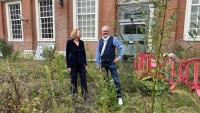 Twee mensen staan in een overwoekerd gebied voor een bakstenen gebouw met hoge ramen. De man draagt een blauw vest en een spijkerbroek, terwijl de vrouw een donker pak draagt. Een deur met het opschrift "Polytechnic Tourism Academy" en rode barrières zijn